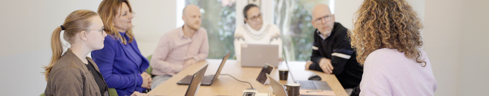In een vergaderruimte op het bestuursbureau is een team van zes mensen in gesprek met elkaar. Op de vergadertafel staan laptops.