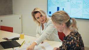 Twee vrouwen zitten aan een tafel; de een laat iets zien aan de ander terwijl ze samen aan beleid werken.
