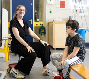 In een schoolklas zit een juf op een kruk tegenover een leerling met een bril die zijn ogen dichtknijpt terwijl hij naar een televisiescherm kijkt. De juf lacht en kijkt in de camera.