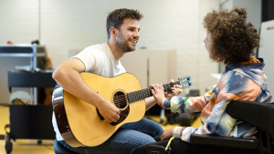 Een muziekleraar speelt op zijn gitaar en kijkt naar een meisje in een rolstoel. Het meisje raakt de gitaar aan.