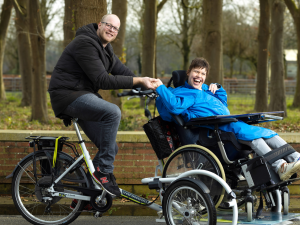 Een mannelijke begeleider zit op een rolstoelfiets met een jonge vrouw. Ze lachen samen, houden elkaars hand vast en zijn buiten, met bomen op de achtergrond.