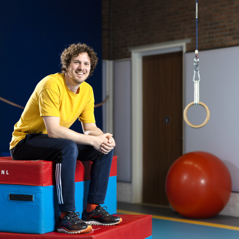 Gymdocent Coen speelman zit in een gymzaal op een sportmat. Er zijn veel contrasterende kleuren te zien in de gymzaal.
