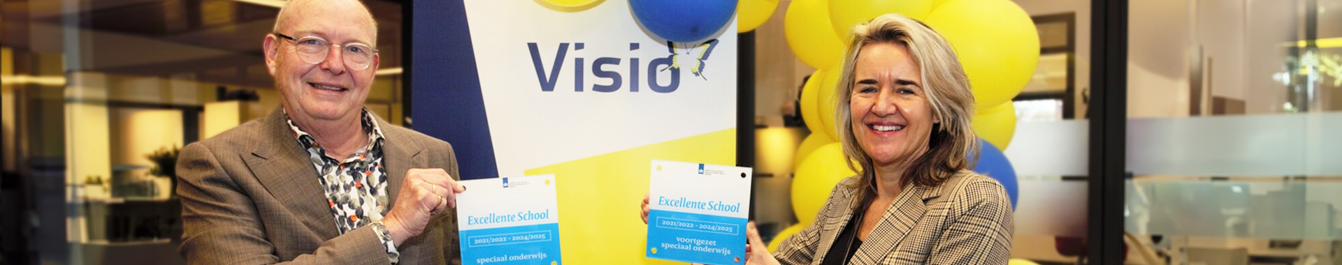 Twee bestuursleden van Visio nemen de onderscheiding voor excellent onderwijs in ontvangst. Ze houden de onderscheiding vast en glimlachen. Er zijn ballonnen op de achtergrond in beeld.