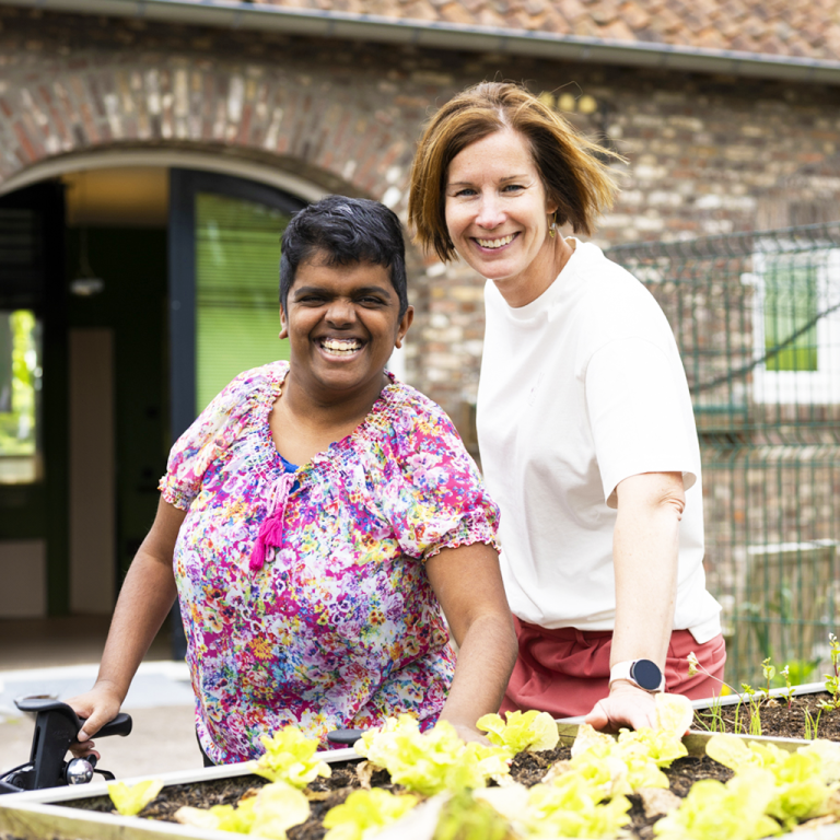 Een Coördinerend cliëntbegeleider en een jonge vrouw met een visuele beperking staan in de tuin bij de planten. Ze lachen en kijken energiek in de camera. De jonge vrouw heeft een rollator vast