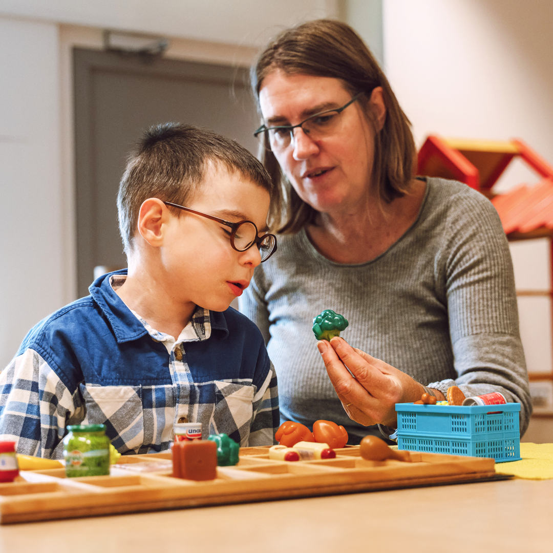Een ergotherapeut zit met een kind aan tafel in een school omgeving. Ze laat hem een tastbare plastic broccoli zien.