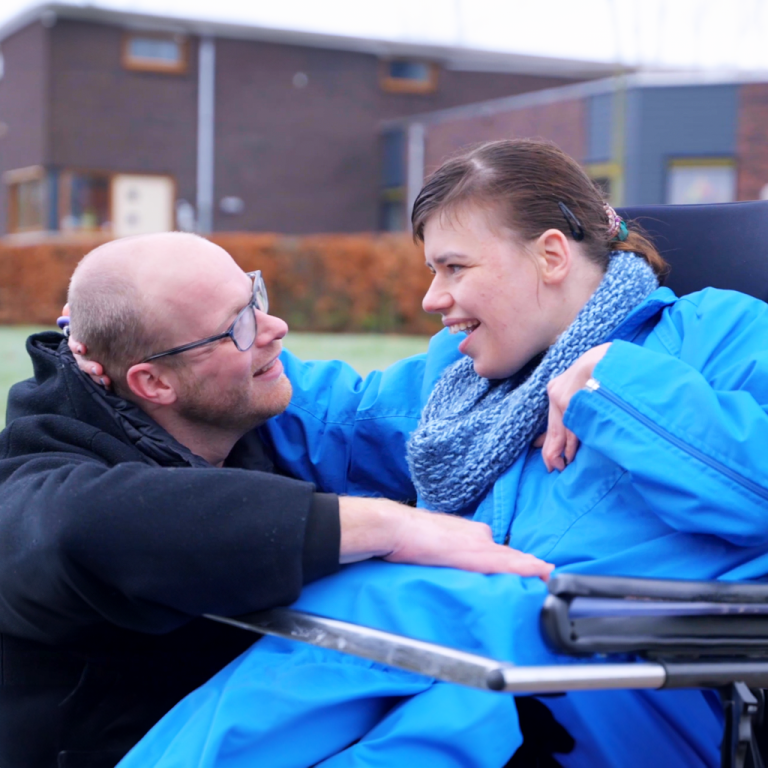 Begeleider Rudie en een jonge vrouw in een rolstoelfiets kijken elkaar lachend aan.