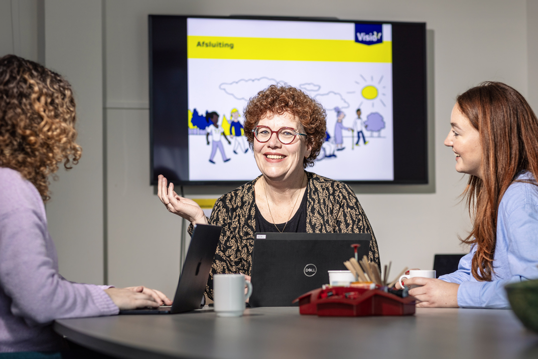 Een adviseur zit aan tafel met twee stagiaires. De adviseur legt iets uit en maakt daarbij een handgebaar.
