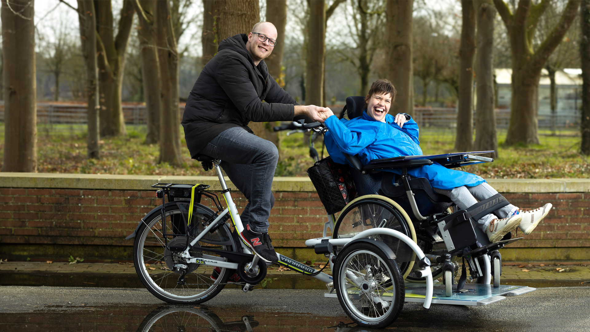 Een mannelijke begeleider zit op een rolstoelfiets met een jonge vrouw. Ze lachen samen, houden elkaars hand vast en zijn buiten, met bomen op de achtergrond.