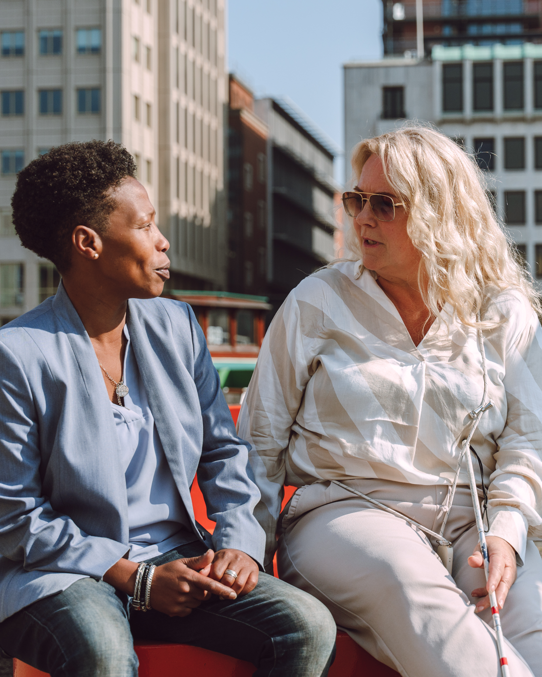 Een slechtziende vrouw met taststok en zonnebril zit buiten in Rotterdam met haar mobiliteitstrainer, een vrouw met een donkere huidskleur.