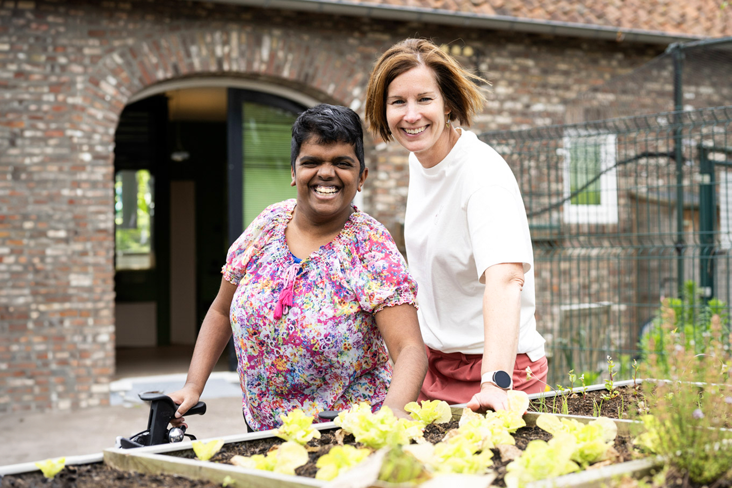 Een Coördinerend cliëntbegeleider en een jonge vrouw met een visuele beperking staan in de tuin bij de planten. Ze lachen en kijken energiek in de camera. De jonge vrouw heeft een rollator vast.