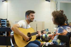 Leerkracht Muziek Een muziek Leerkracht speelt gitaar voor een jonge vrouw in een rolstoel. Zij raakt de hals van de gitaar aan.