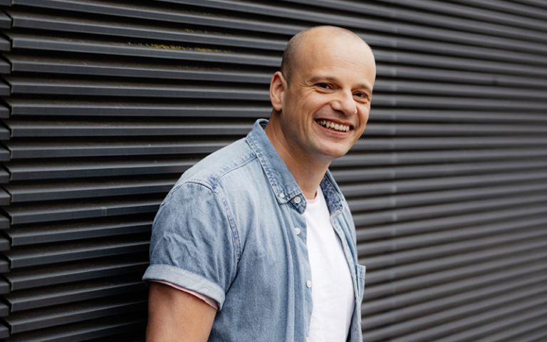 Begeleider Andy leunt tegen een buitenmuur en kijk lachend.