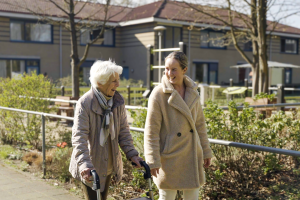 Een medewerker Verzoring loopt buiten op het terrein van Visio Vught met een oudere dame.