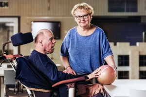 Een man in een rolstoel doet een oefening met een fysiotherapeut. De fysiotherapeut, een vrouw met kort blond haar en een blauwe trui, helpt hem terwijl hij een oefenbal vasthoudt op tafel.