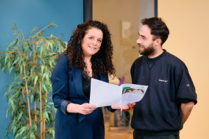 Vriendelijke vrouwelijke manager met zwarte krullen legt iets uit op een document aan een bebaarde medewerker in een kantoorruimte, terwijl ze glimlachend in de camera kijkt.