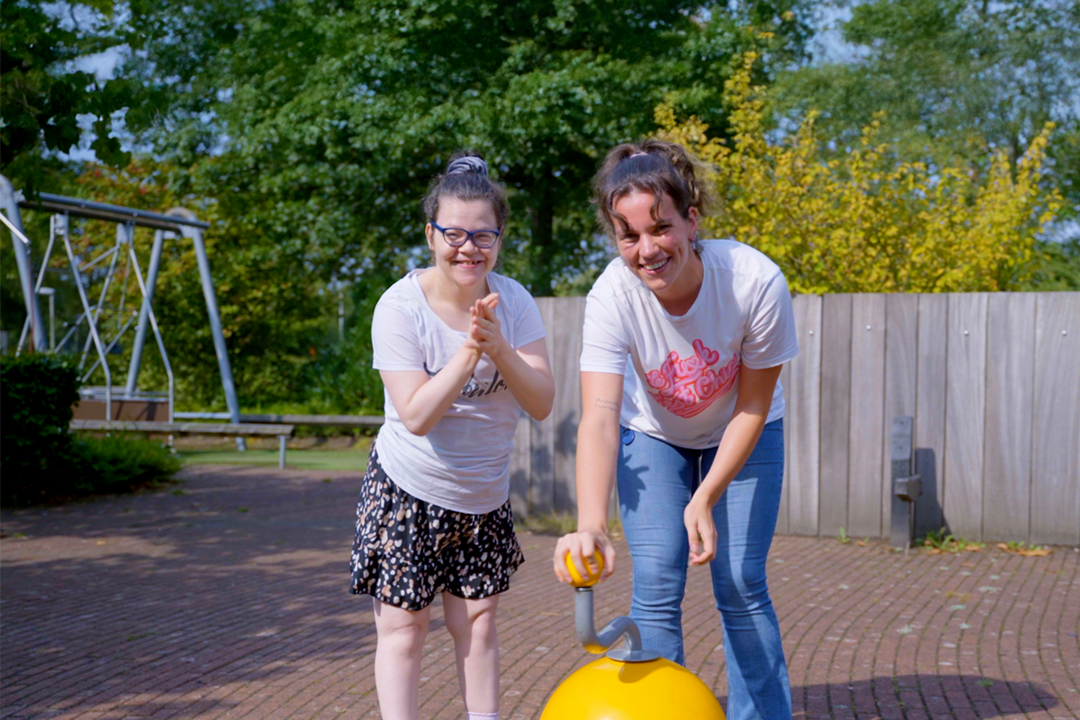 Een jonge vakantie medewerker maakt muziek in de buitenspeelplaats, samen met een jonge vrouwelijke cliënt, ze kijken vrolijk en het meisje klapt in haar handen.
