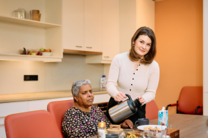 Een jonge medewerker staat in een keuken en schenkt koffie aan een client. De client zit aan tafel.