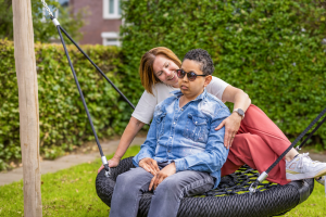 Een begeleider en een blinde jongen zitten samen buiten op een grote schommel.