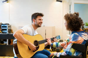 Een muziek Leerkracht speelt gitaar voor een jonge vrouw in een rolstoel. Zij raakt de hals van de gitaar aan.